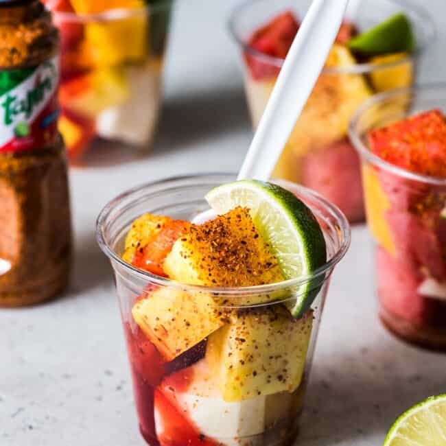 Mexican Fruit Cups on a table topped with chile lime seasoning, chamoy and a lime wedge.