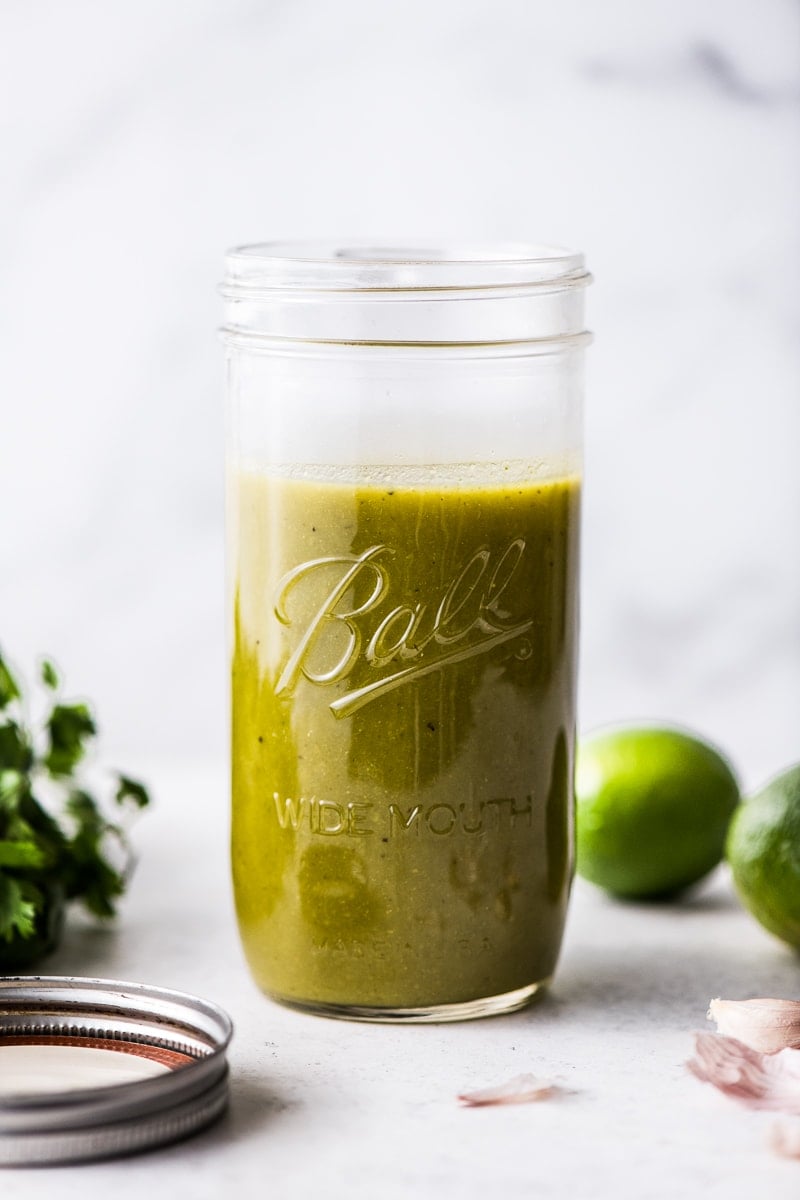 A jar of green enchilada sauce next to limes, garlic cloves and cilantro.