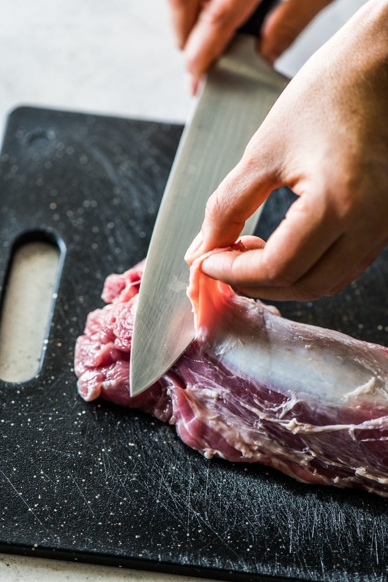 A knife and hand removing silver skin from a pork tenderloin.