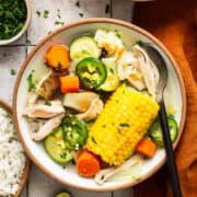 Caldo de pollo (Mexican chicken soup) in a bowl alongside white rice, cilantro, limes, and tabasco.