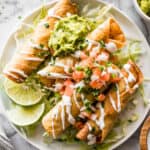 Chicken taquitos on a plate garnished with Mexican crema, lettuce, guacamole, and pico de gallo.