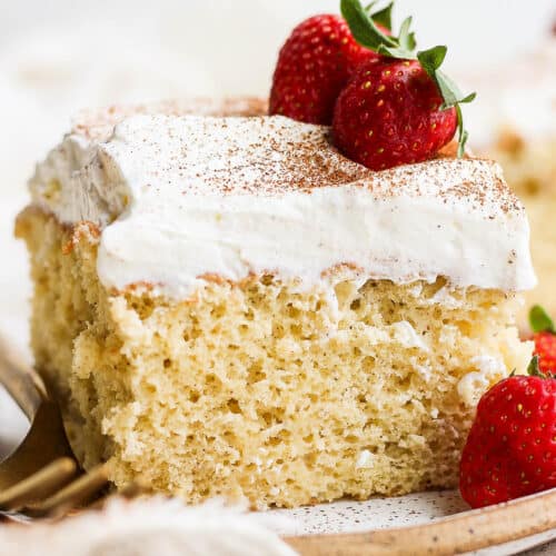 A slice of tres leches cake on a plate topped with whipped cream and fresh strawberries.