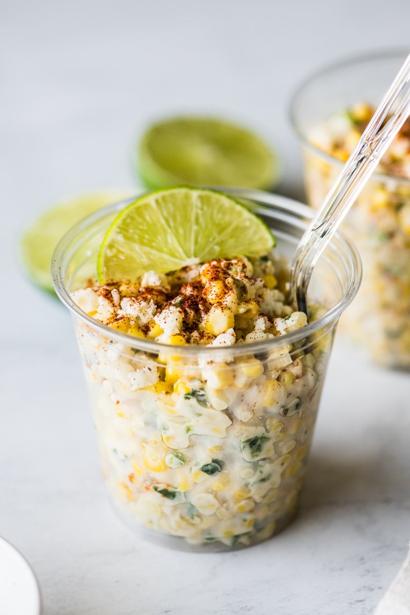 A clear plastic cup filled with esquites and a plastic spoon in it.