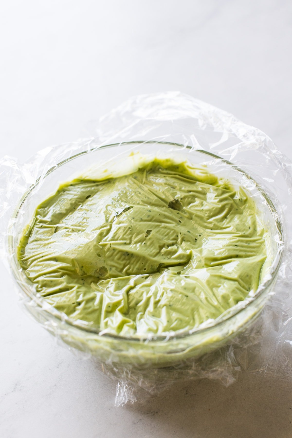 A bowl of avocado crema with clear plastic wrap on top.