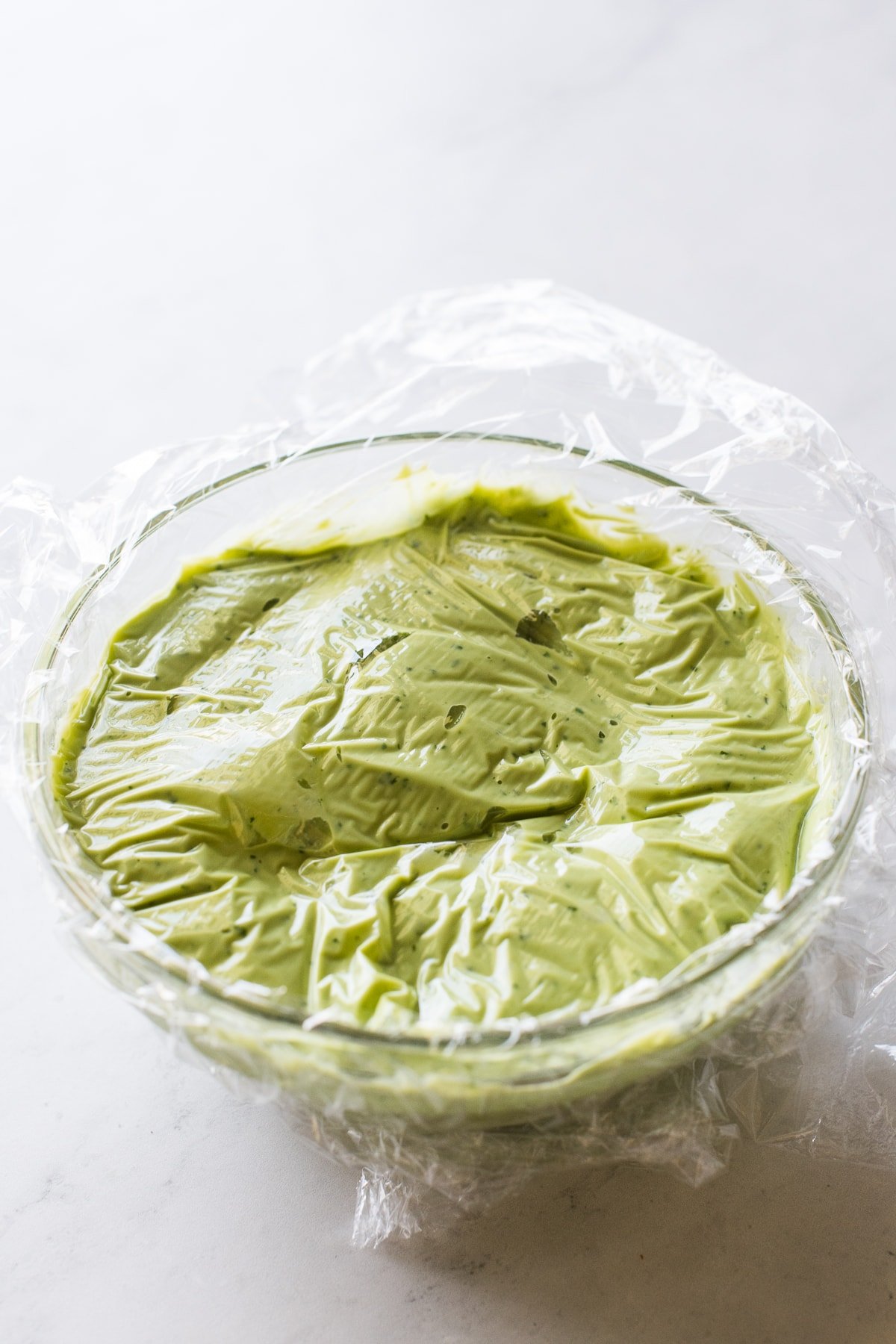 A bowl of avocado crema with clear plastic wrap on top.