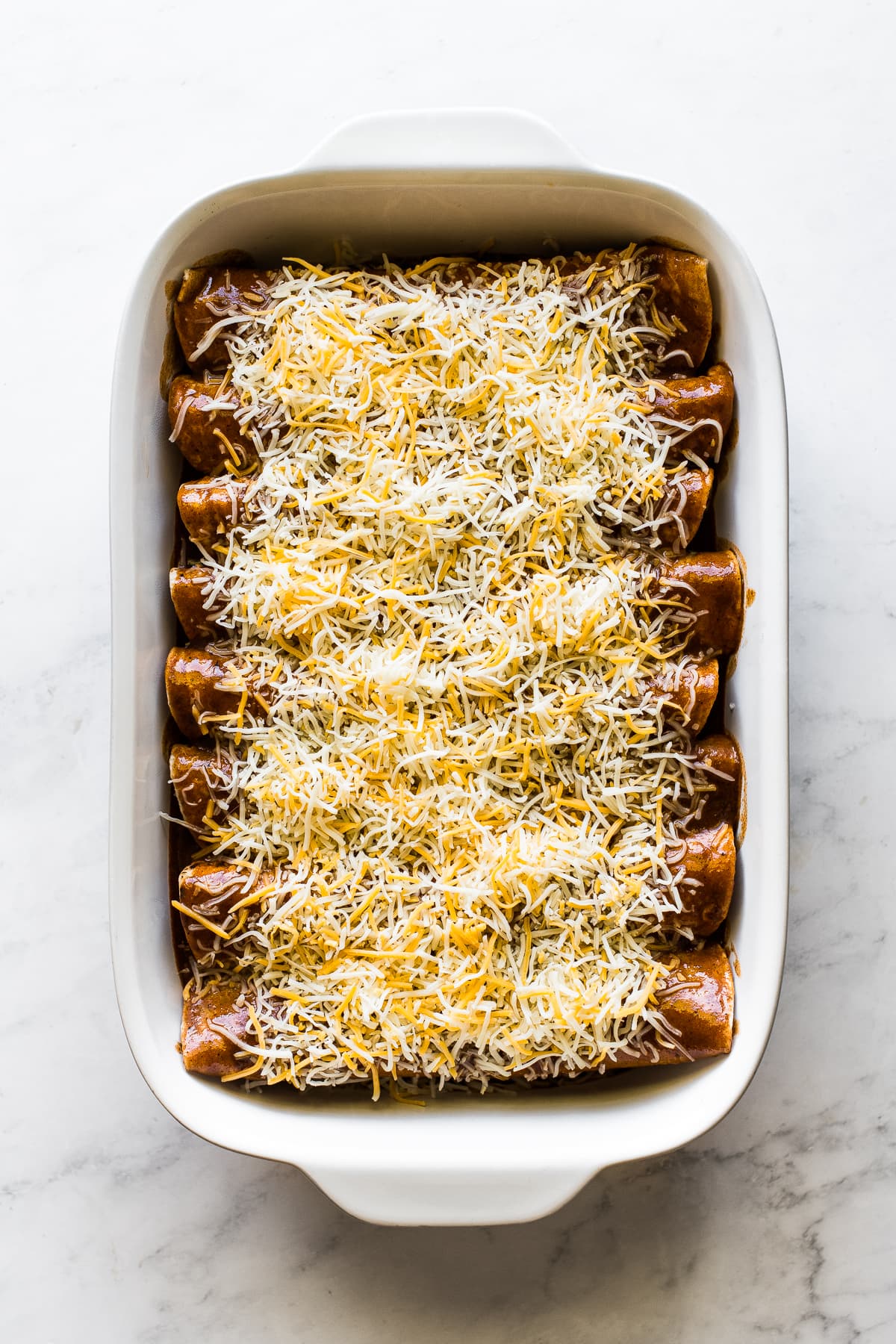 Enchiladas ready to be baked in a pan topped with enchilada sauce and shredded cheese.