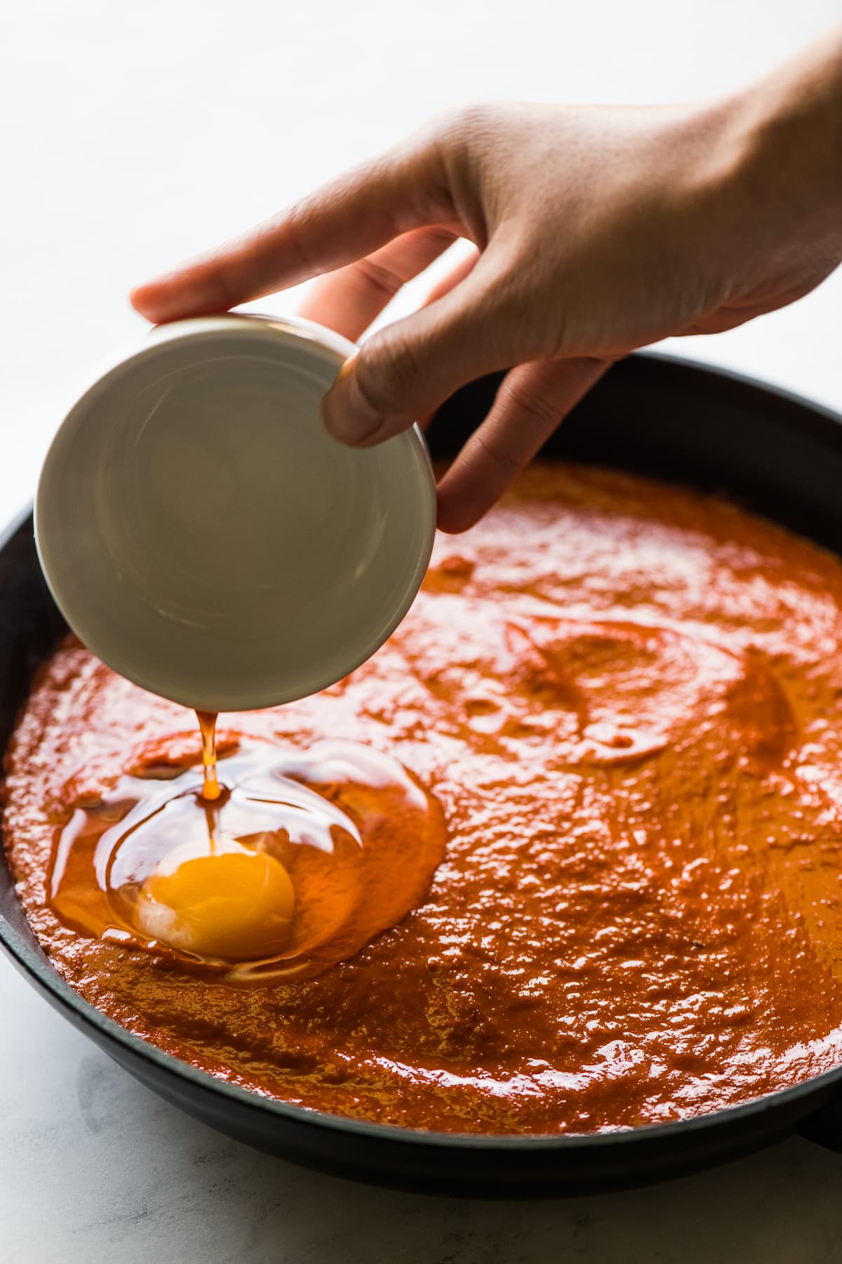 An egg being poured into a skillet with salsa for huevos ahogados.