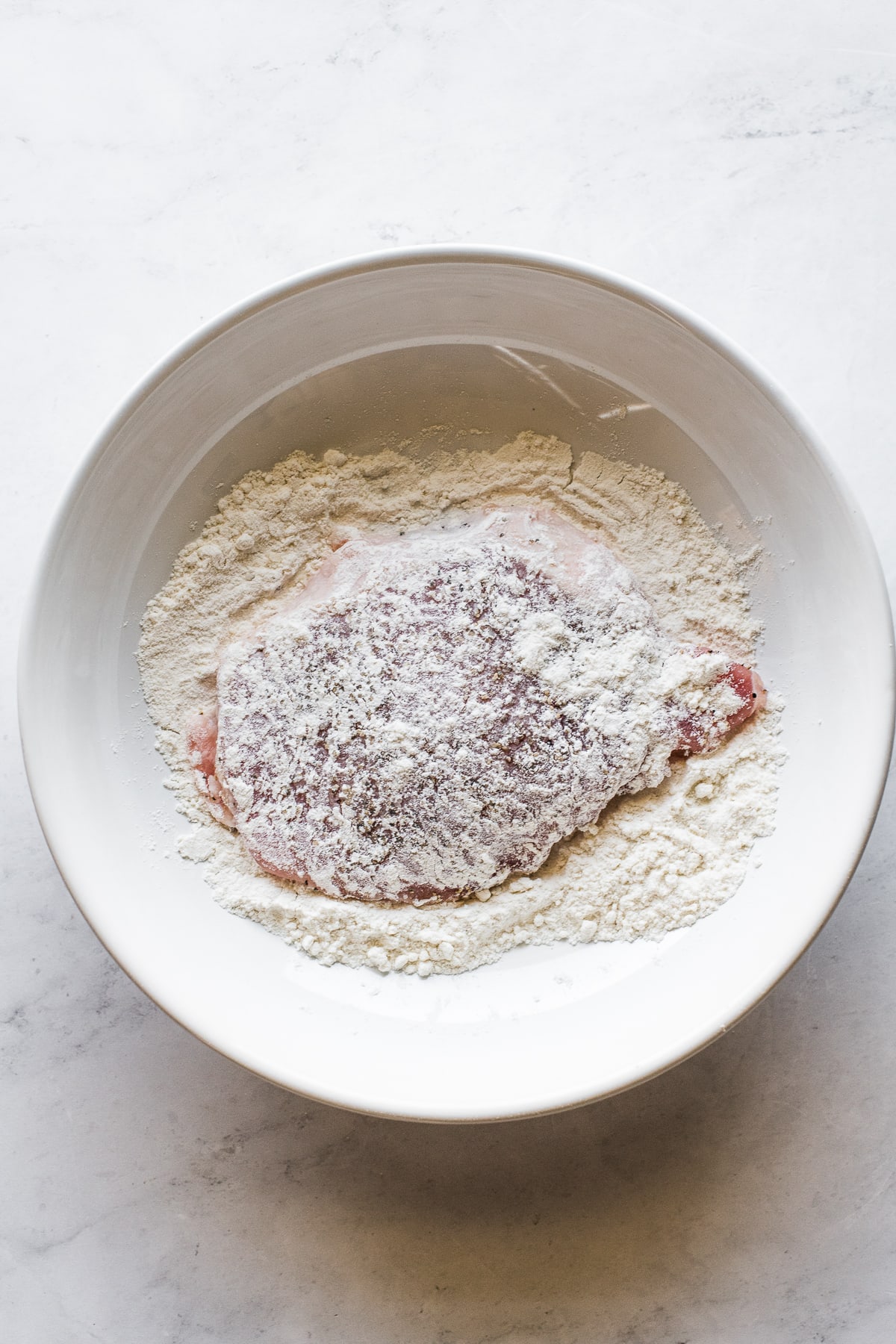 A pork cutlet in a shallow bowl filled with all-purpose flour.