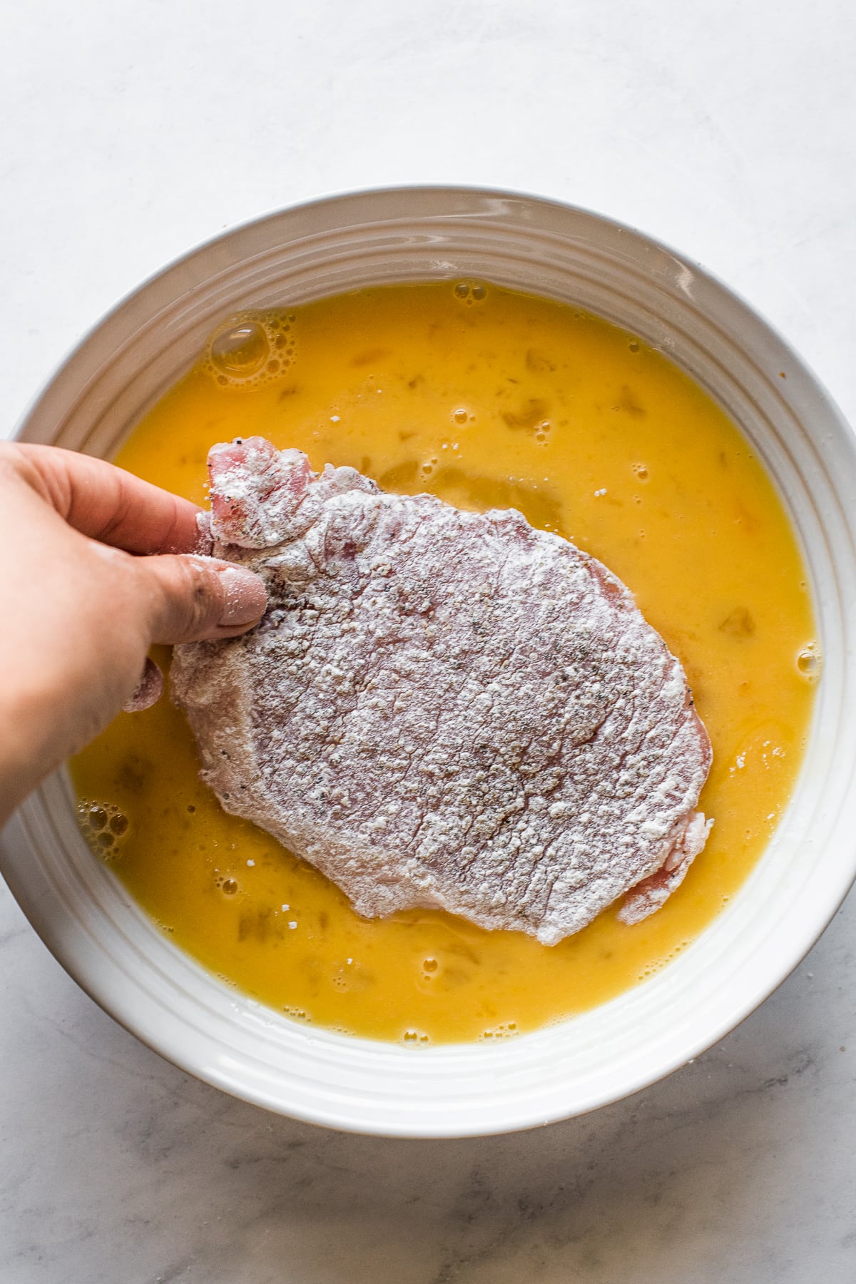 A pork cutlet being dipped in a bowl of beaten eggs.