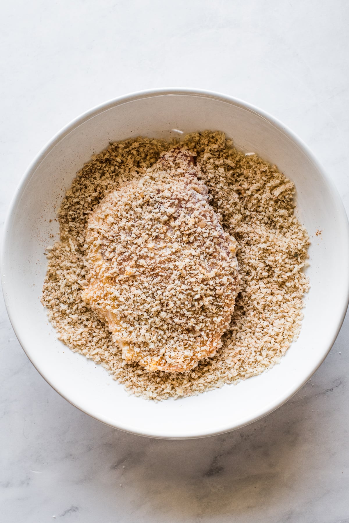 A pork chop in a bowl full of seasoned panko breadcrumbs.