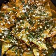 Chilaquiles verdes, aka green chilaquiles, in a skillet topped with ciantro, onion, and cotija cheese.