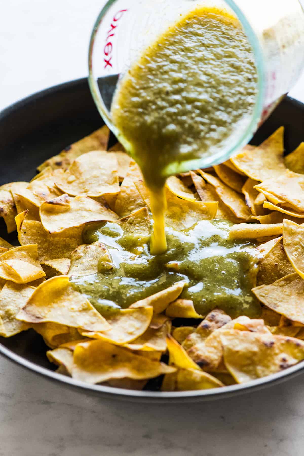 Fried corn tortillas in a skillet with salsa verde being poured on top of them for green chilaquiles.