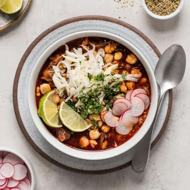 Red posole in a bowl topped with cabbage, radishes, cilantro, and limes.