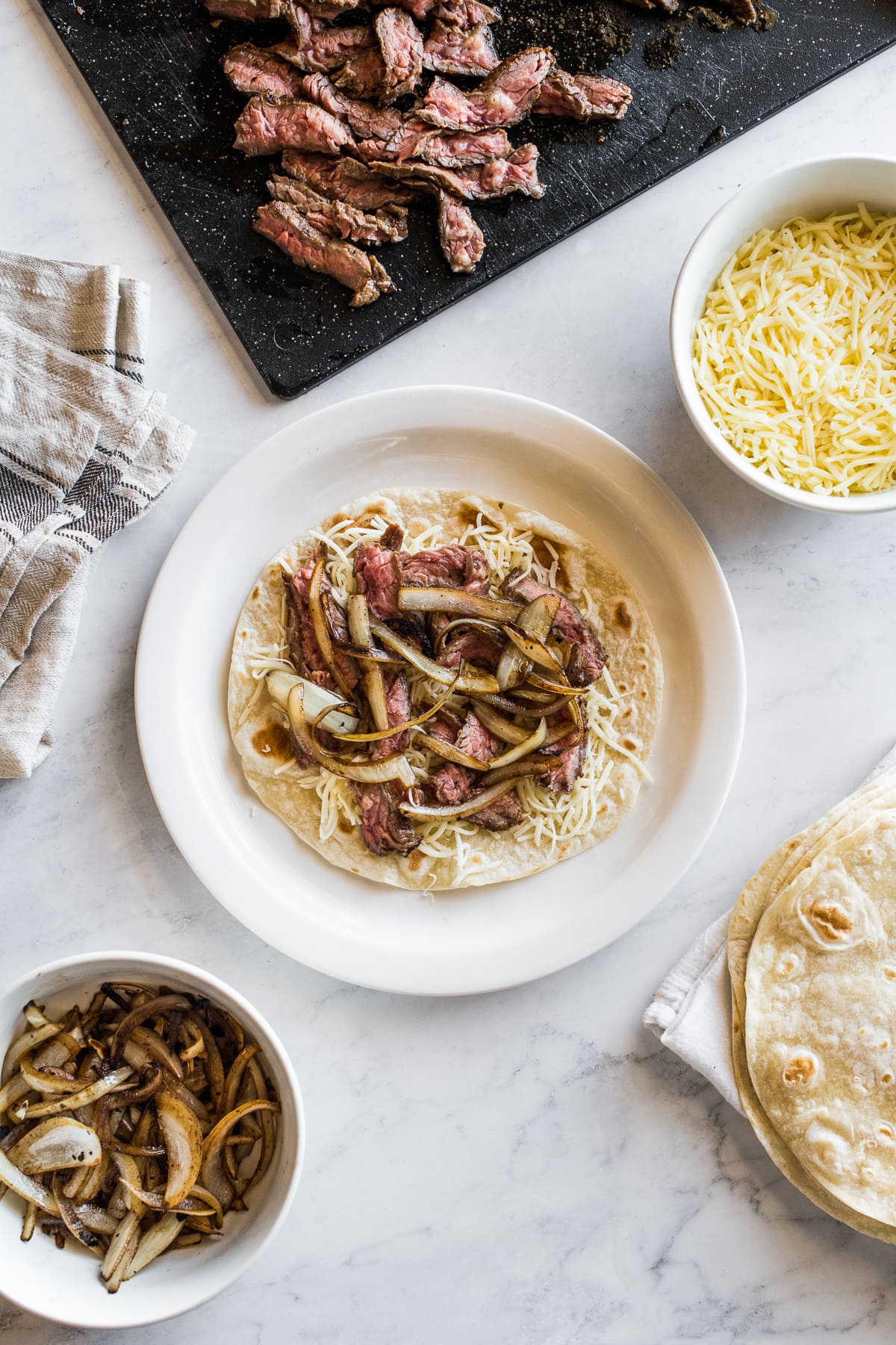 A steak quesadilla being assembled with sliced steak, shredded cheese, and sauteed onions.