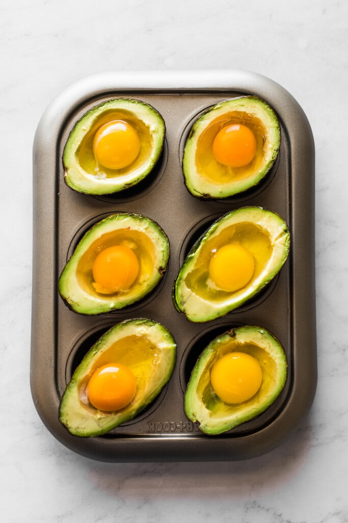 Avocado halves on a muffin tin with gently cracked eggs inside each well.