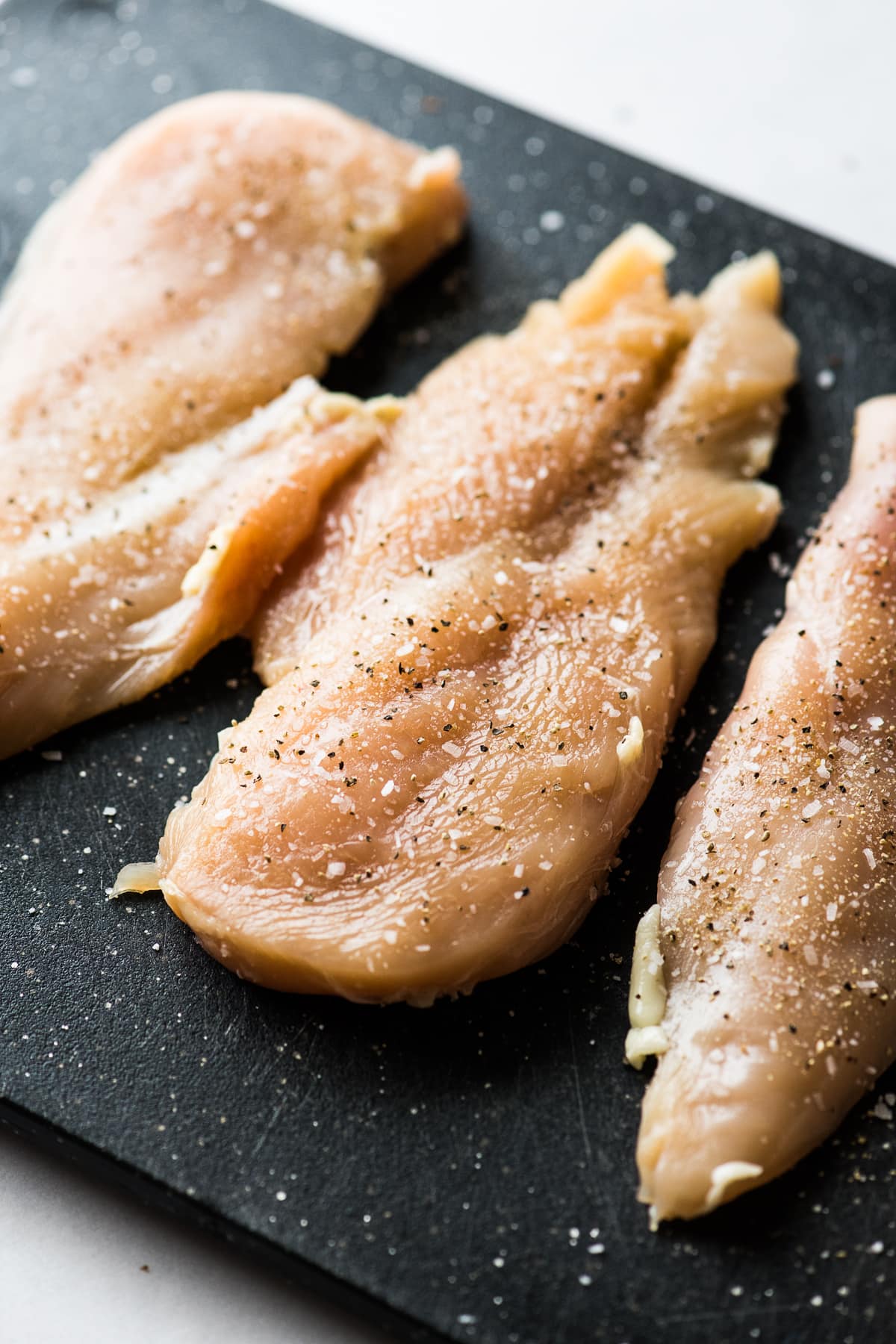 Sliced chicken breast on a cutting board sprinkled with salt and black pepper.