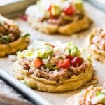 Sopes on a baking sheet topped with refried beans, lettuce, and tomatoes.