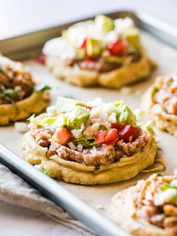 Mexican sopes recipe on a baking sheet topped with refried beans, lettuce, and tomatoes.