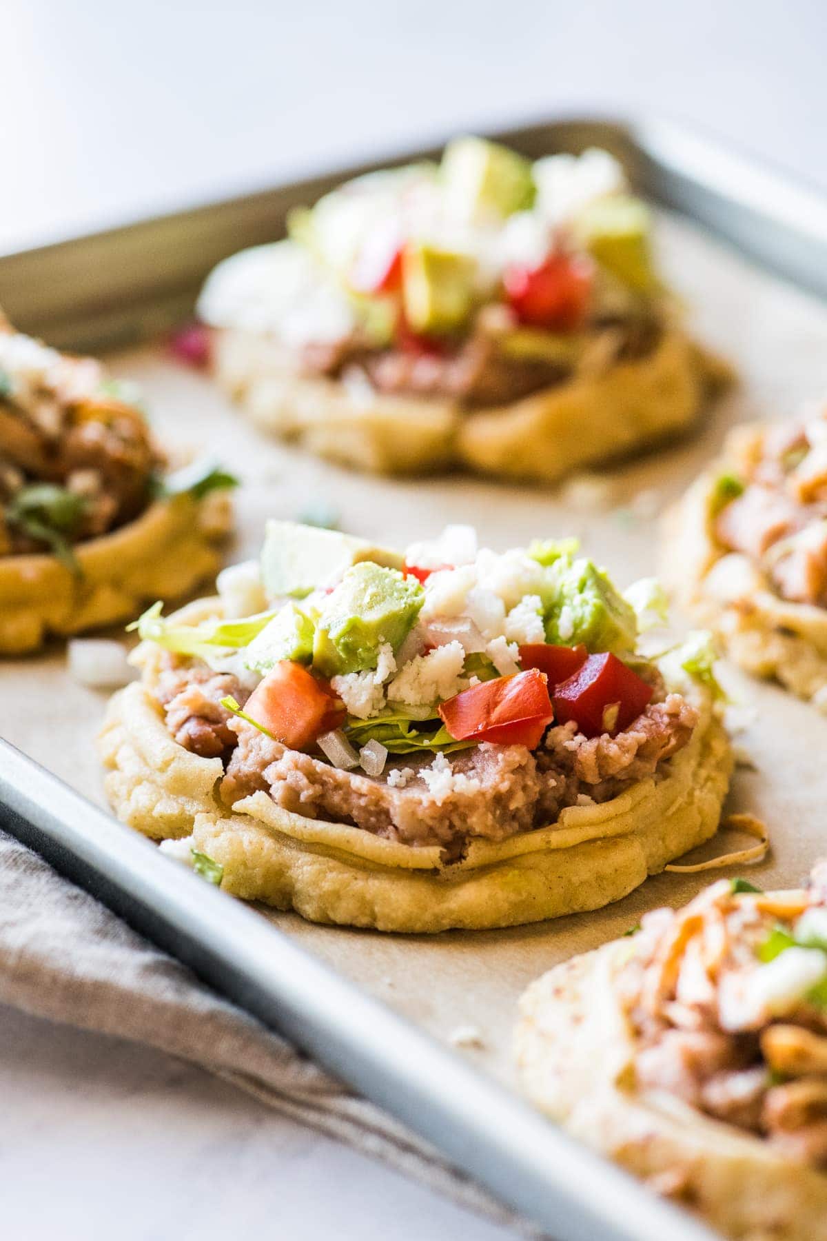 Sopes on a baking sheet topped with refried beans, lettuce, and tomatoes.
