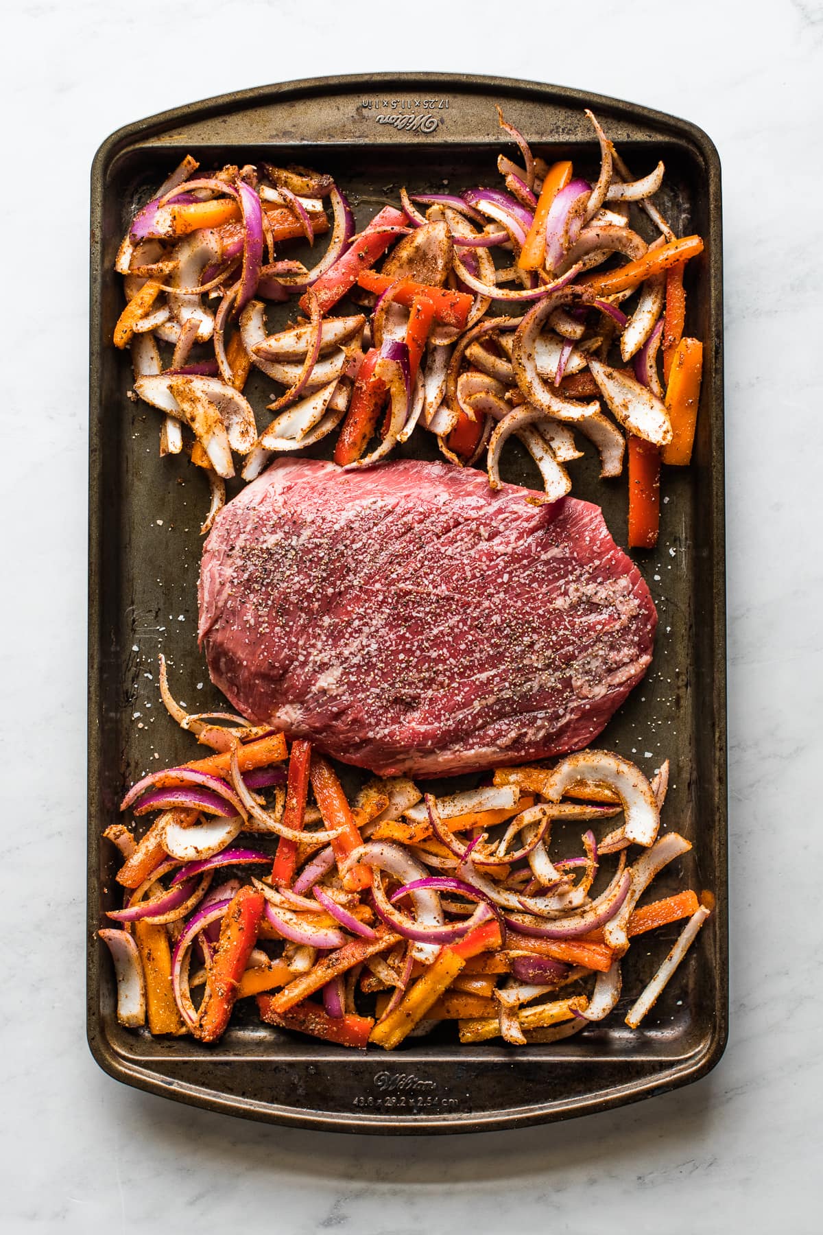 A sheet pan with flank steak and sliced peppers and onions.