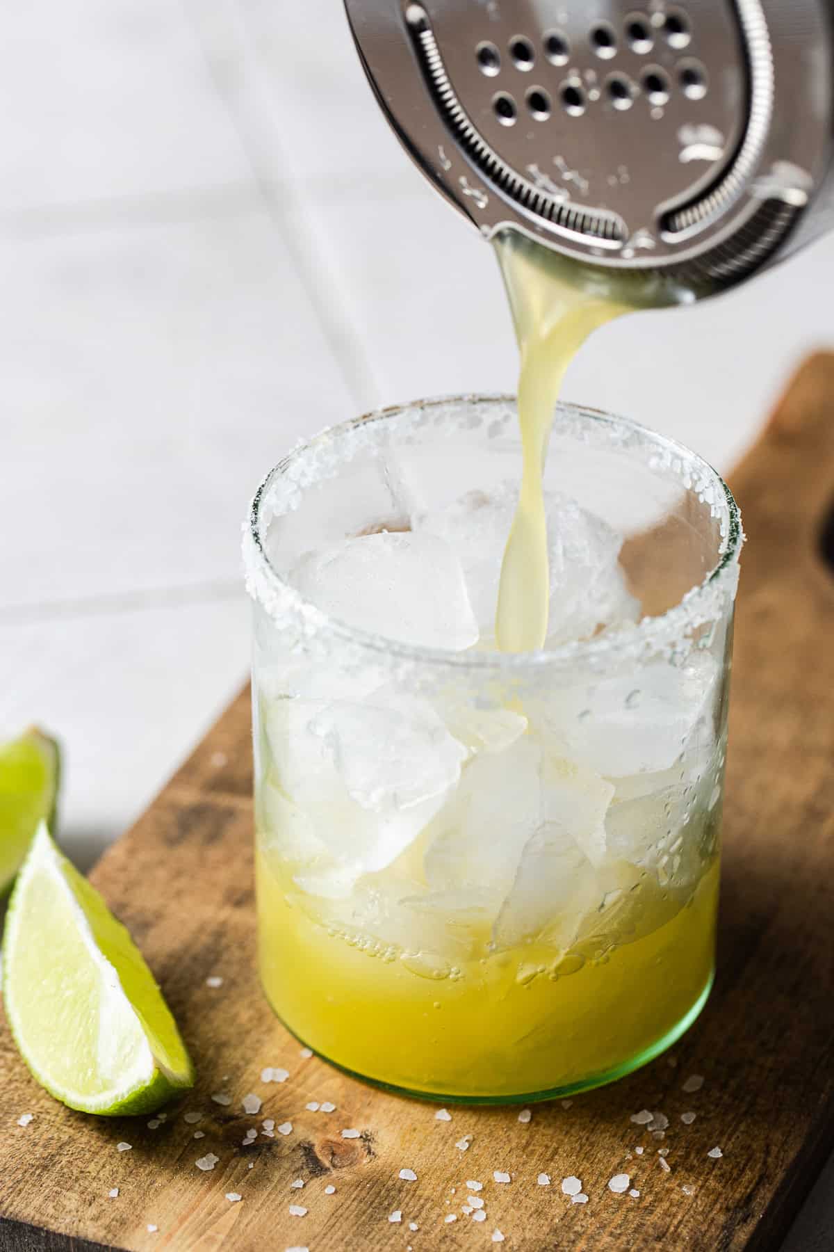 A margarita mocktail being poured from a cocktail shaker into a rimmed and iced glass.