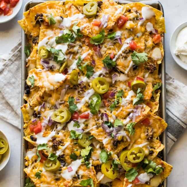 Sheet pan nachos topped with sour cream, pickled jalapenos, tomatoes, and cilantro.