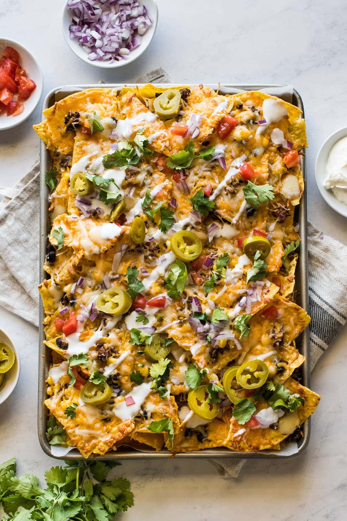 Sheet pan nachos topped with sour cream, pickled jalapenos, tomatoes, and cilantro.