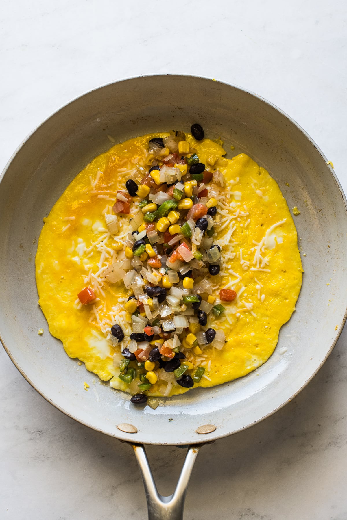 An omelet in a skillet filled with cheese, beans, corn, onions, peppers, and tomatoes.