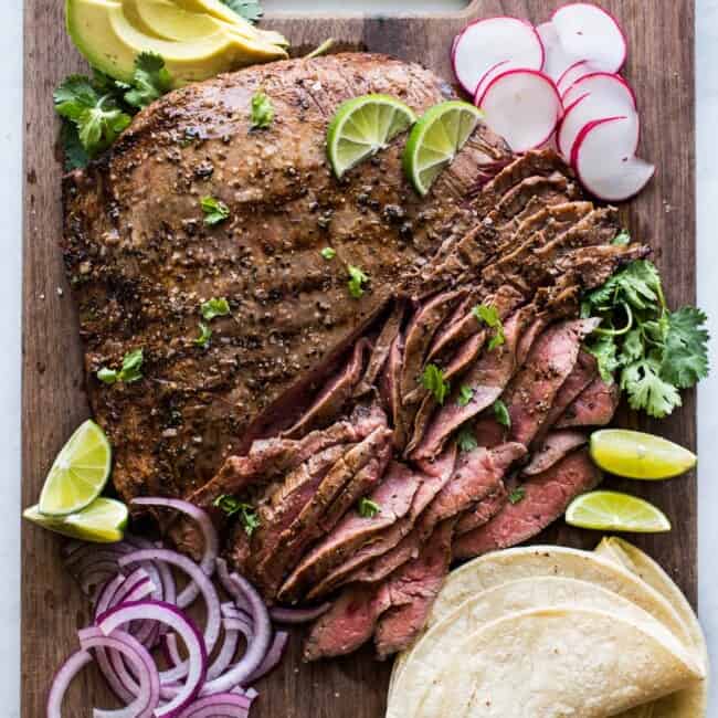 Thinly sliced carne asada on a cutting board with cilantro, corn tortillas, limes, avocados, and radishes.