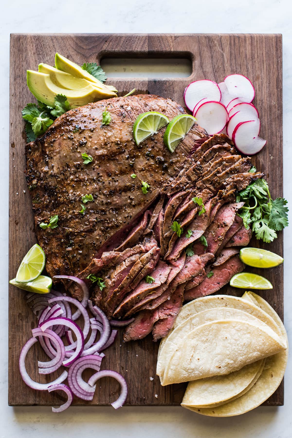 Thinly sliced carne asada on a cutting board with cilantro, corn tortillas, limes, avocados, and radishes.