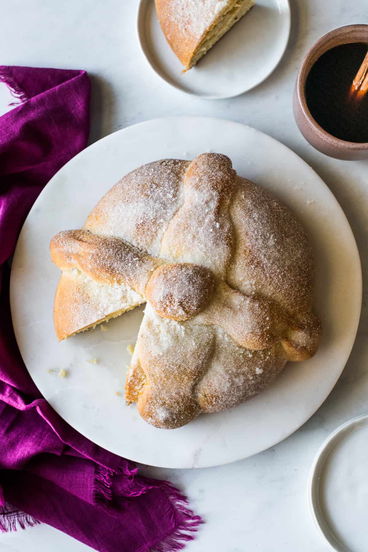 A loaf of Pan de Muerto bread garnished with sugar on top and a slice cut out ready to eat.