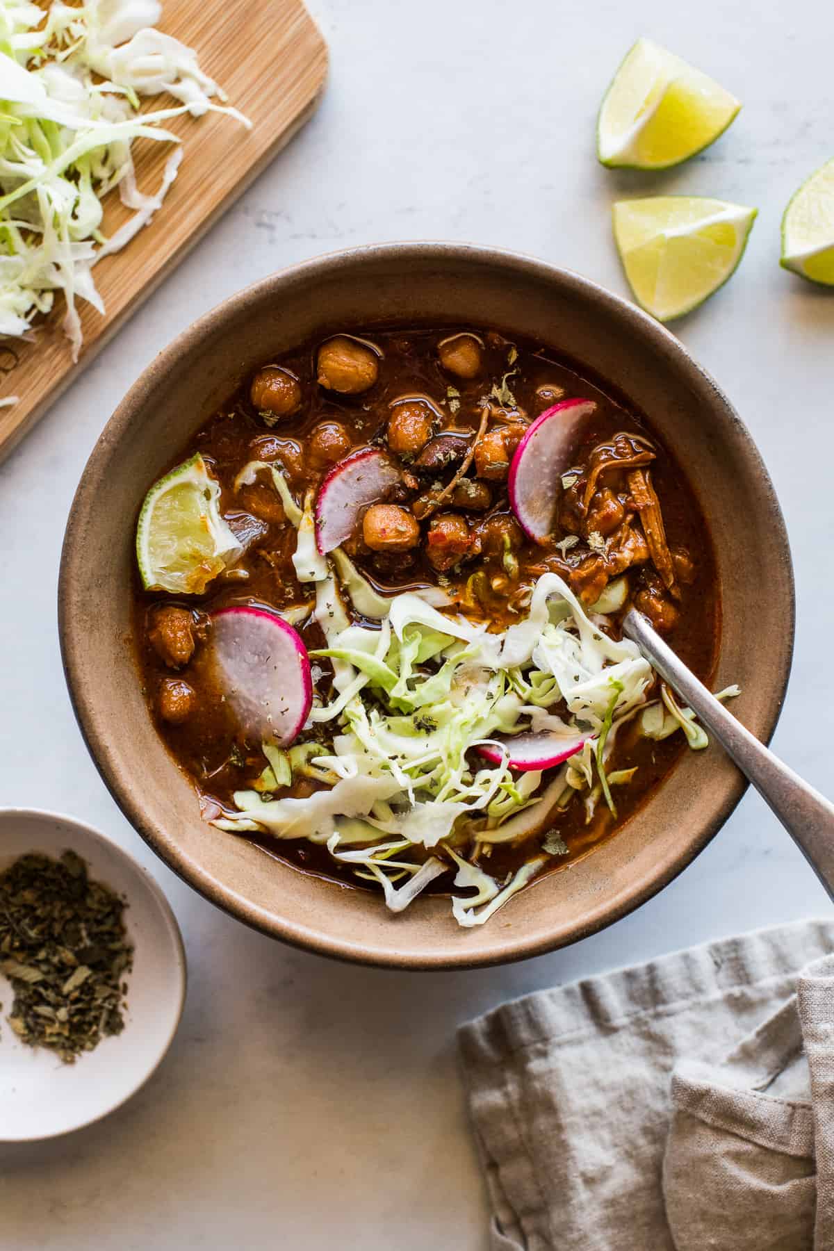 A bowl of pozole rojo garnished with thinly sliced radishes, shredded cabbage, lime wedges and dried Mexican oregano.