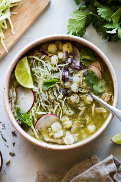 Pozole verde in a bowl topped with shredded cabbage, sliced radishes, cilantro, and lime juice.