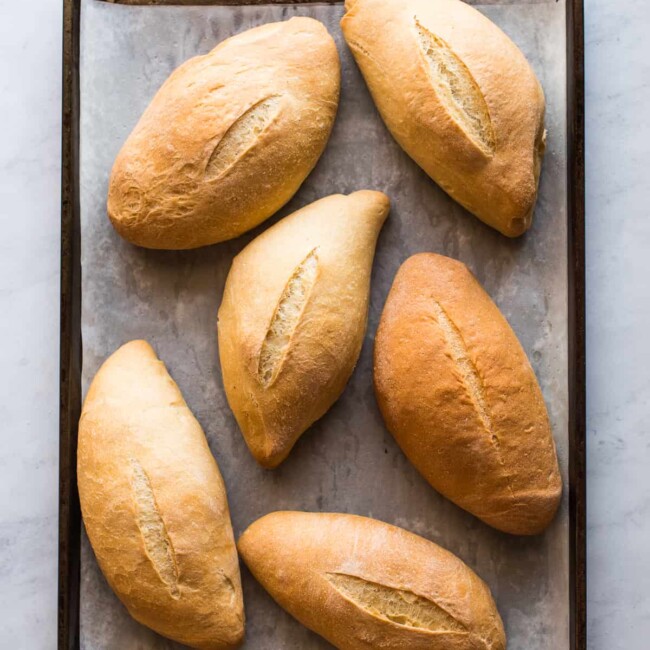 Six bolillo breads on a baking sheet ready to eat.