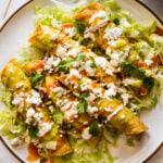 Flautas on a plate topped with crema, hot sauce, salsa verde, lettuce, cilantro, and cotija cheese.