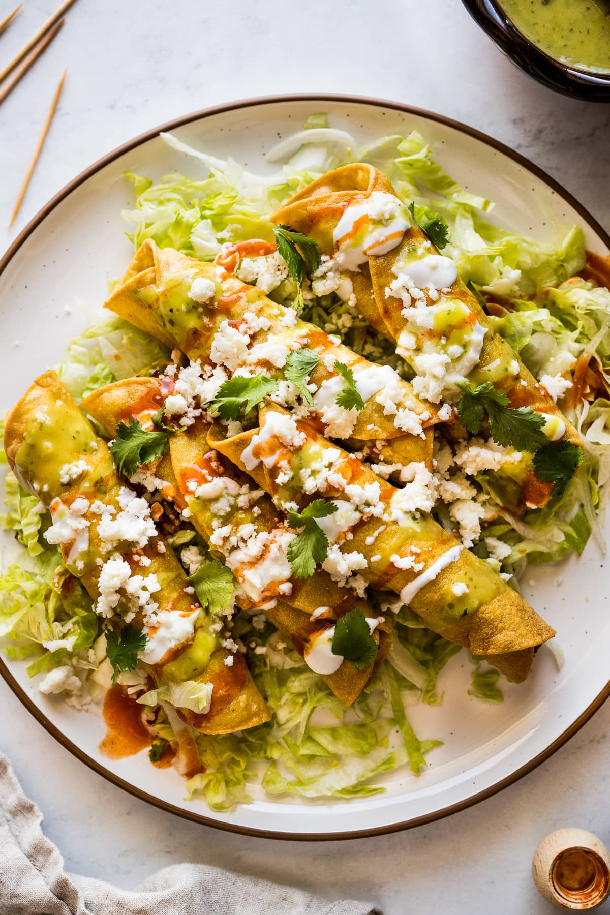 Flautas on a plate topped with crema, hot sauce, salsa verde, lettuce, cilantro, and cotija cheese.