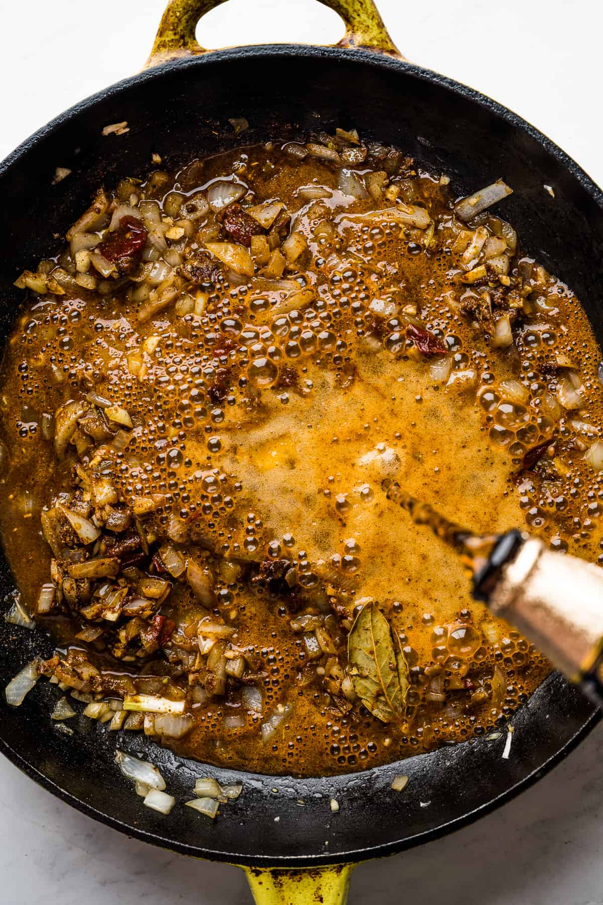 Beer being added to the skillet with cooked onions, garlic, apple cider vinegar, lime juice, spices, and chipotle peppers.