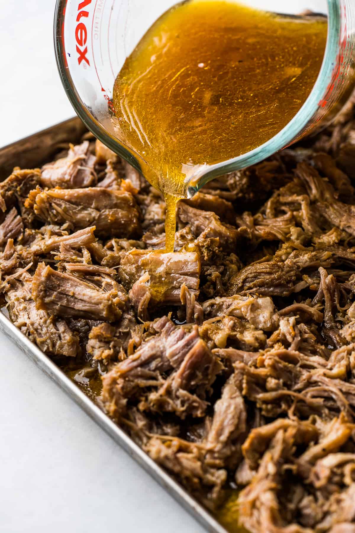 The cooking liquid from carnitas being poured on top of shredded carnitas on a baking sheet.