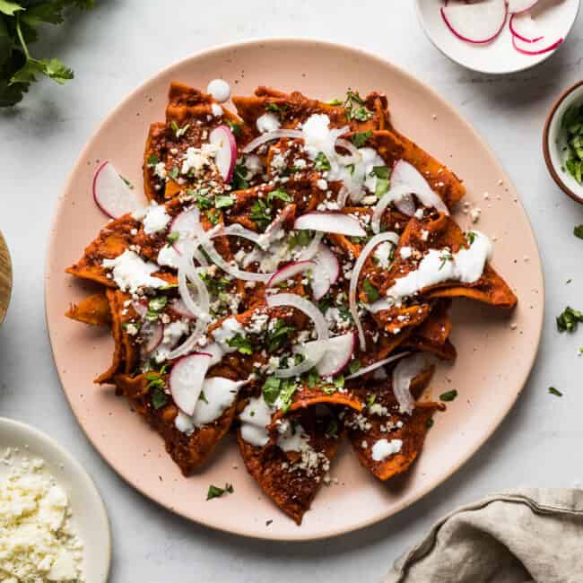 Chilaquiles on a plate with red sauce and topped with Mexican crema, onions, cotija cheese, and cilantro.