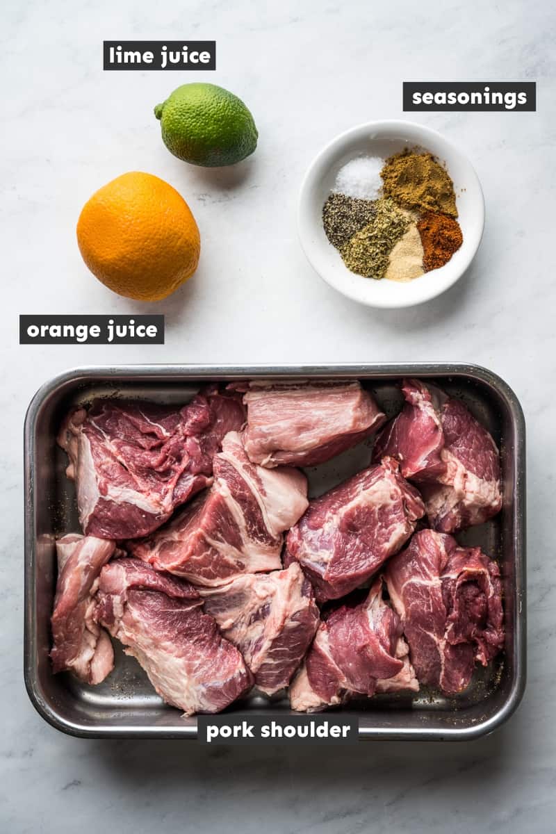 Ingredients for carnitas recipe with all spices in a bowl, orange, lime, and raw pork shoulder in a dish. 