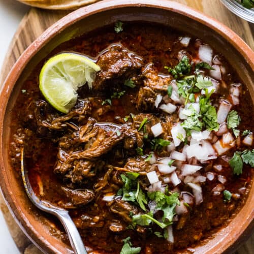 A bowl of beef birria topped with chopped cilantro, onions, and a lime wedge.