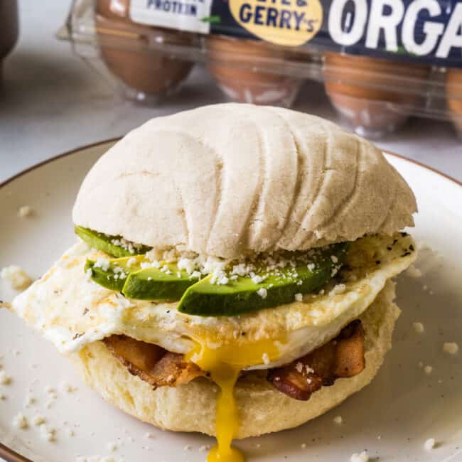 A concha breakfast sandwich on a plate made from bacon, a fried egg, avocado, cotija cheese, and a maple conchas.