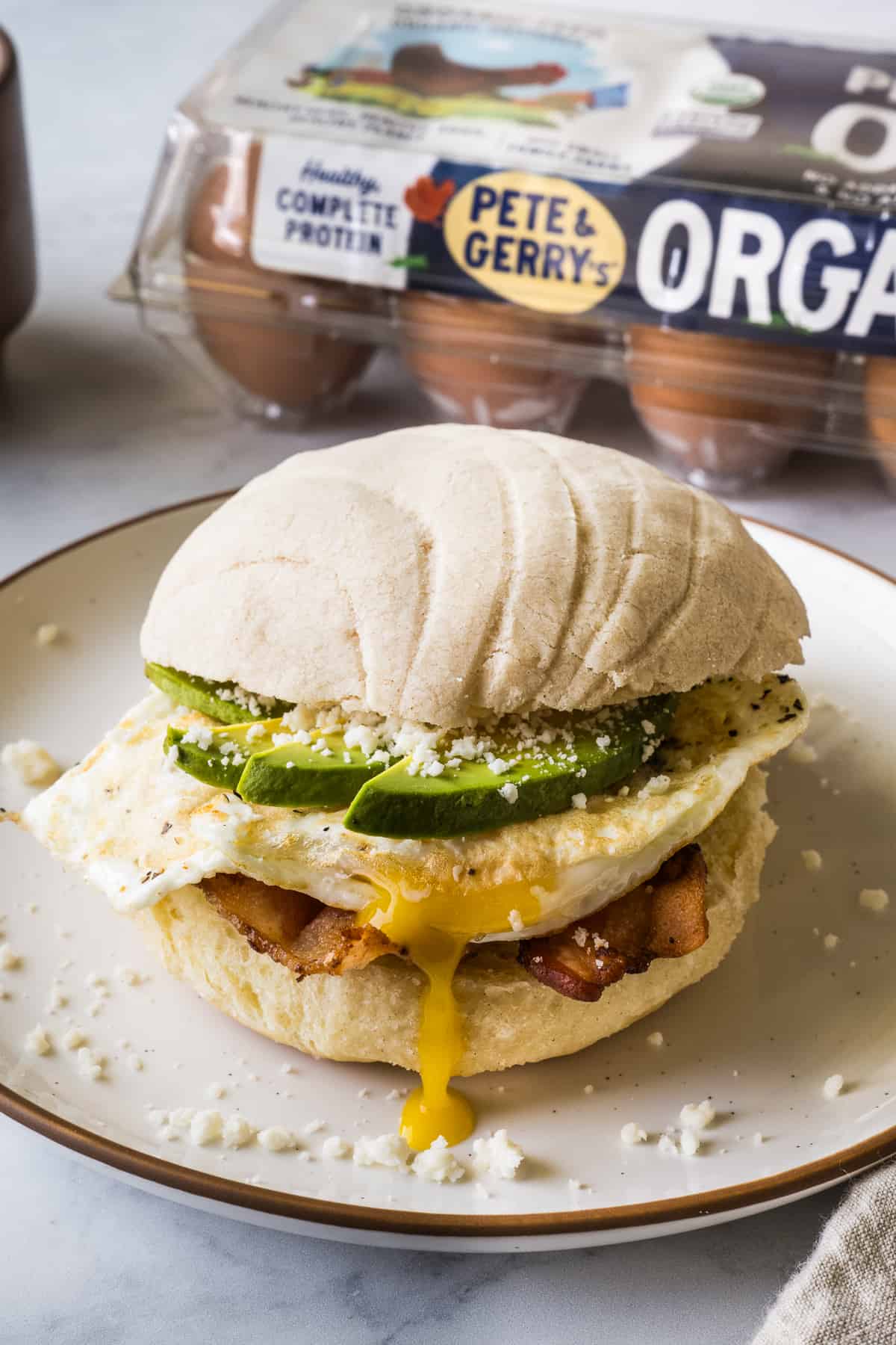 A concha breakfast sandwich on a plate made from bacon, a fried egg, avocado, cotija cheese, and a maple conchas.