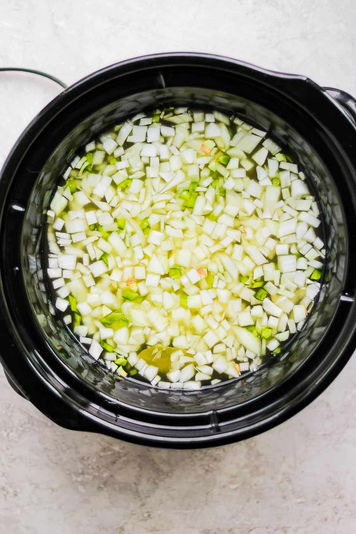 A crock pot filled with onions, peppers, broth, and pinto beans.