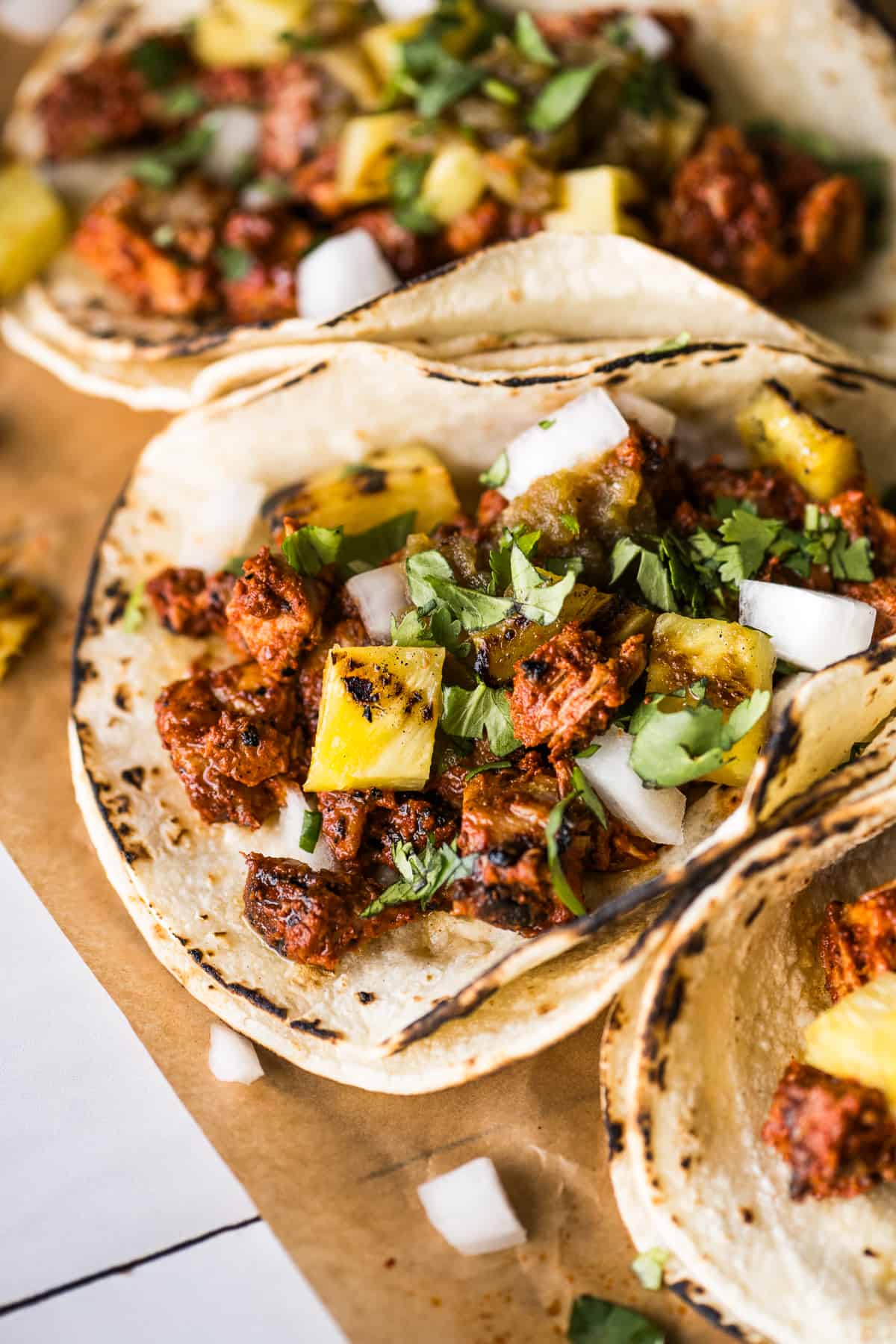 Tacos al pastor in warm corn tortillas topped with grilled pineapple, cilantro, diced white onion, and salsa verde.