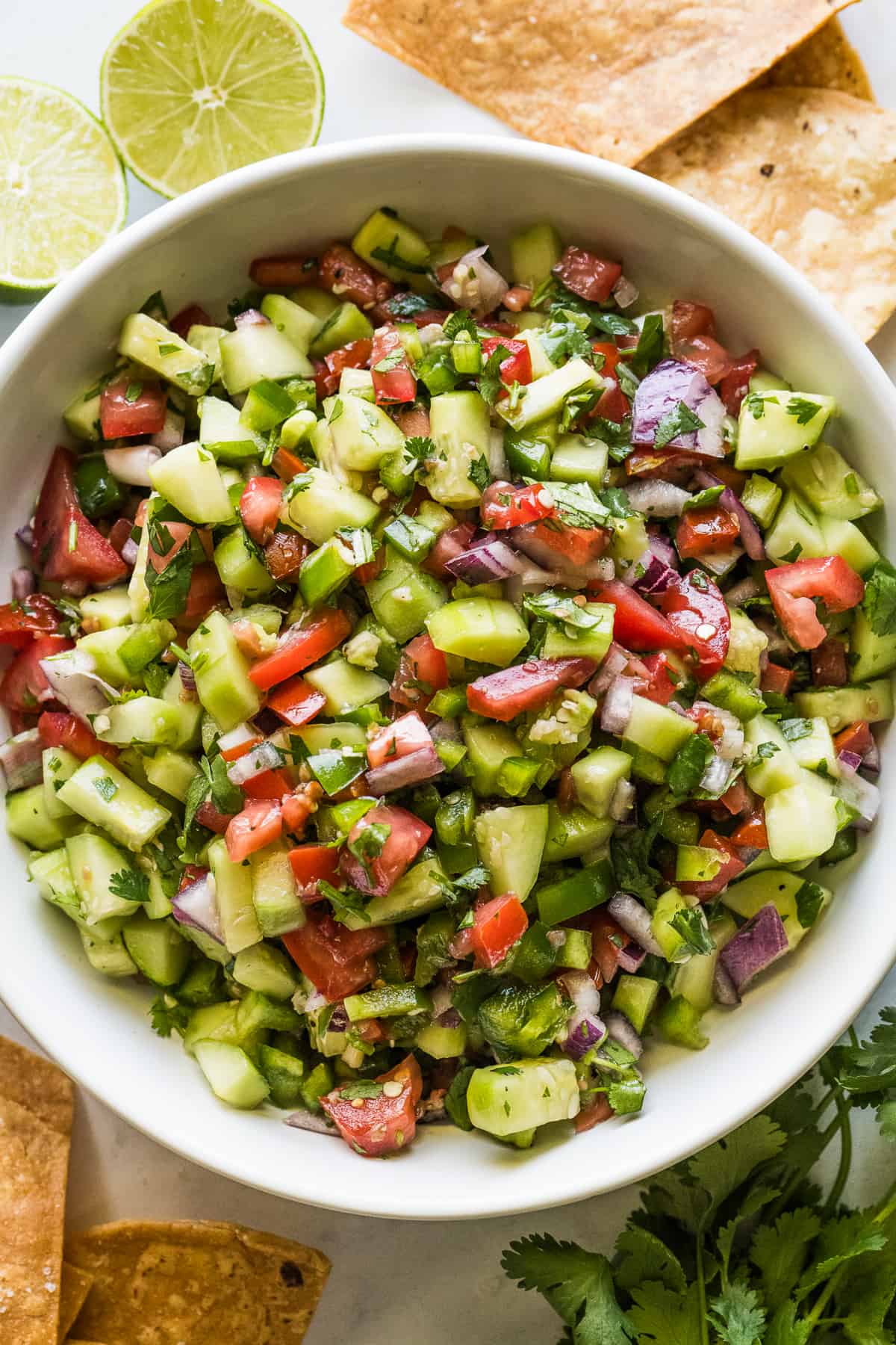 Cucumber Salsa in a bowl with tortilla chips, lime wedges, and cilantro on the side.