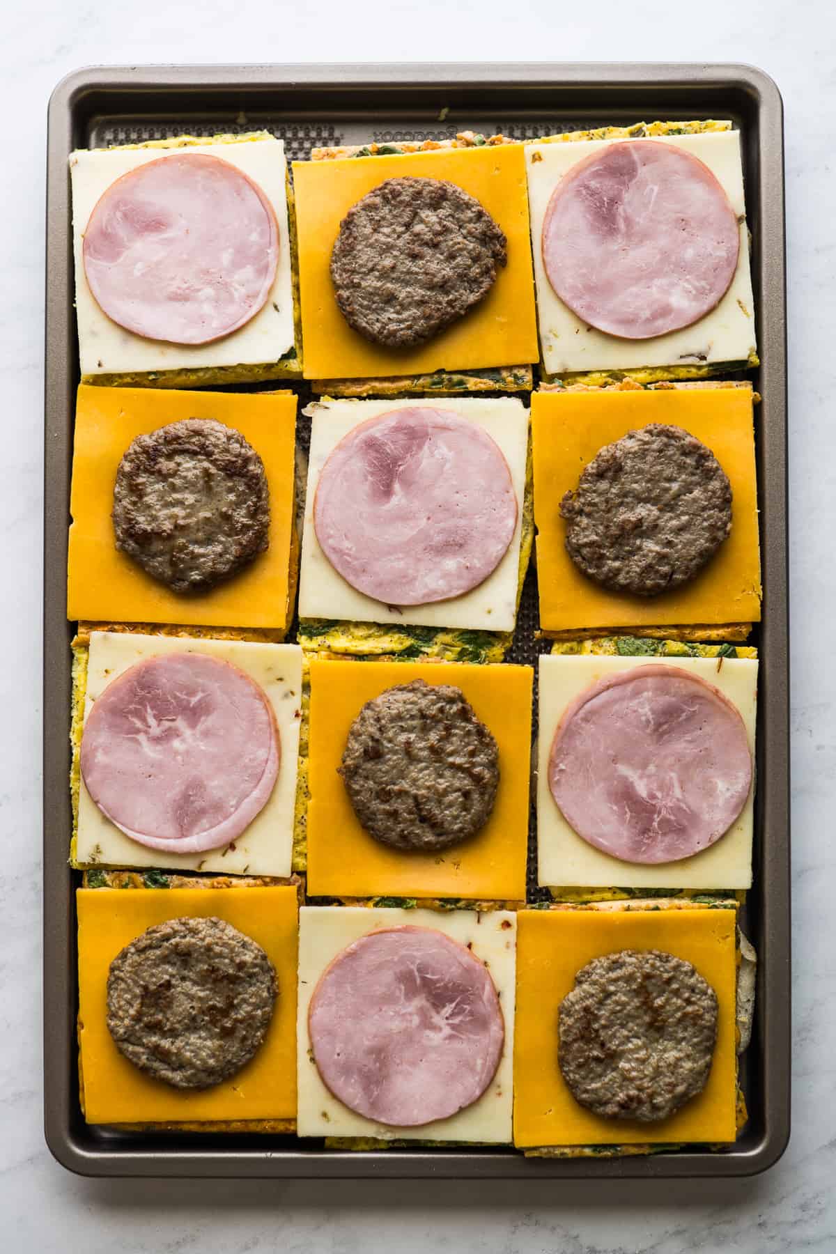 12 English muffins on a sheet pan topped with slices of baked eggs, cheese, and Canadian bacon and sausage.