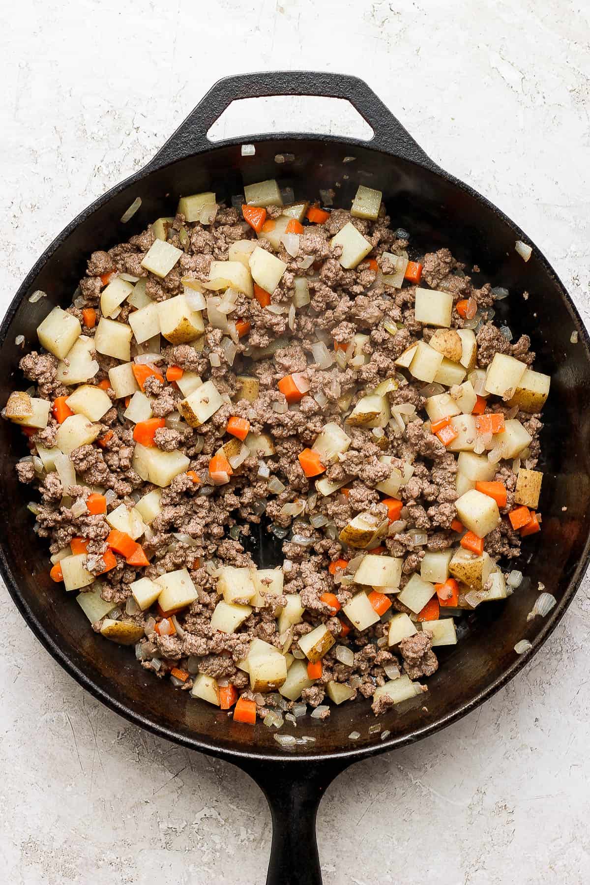 Ground beef, onions, diced potatoes, and diced carrots cooking in a skillet.