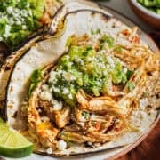 A crockpot shredded chicken taco on a plate with guacamole, cilantro, cotija cheese, and a lime wedge.