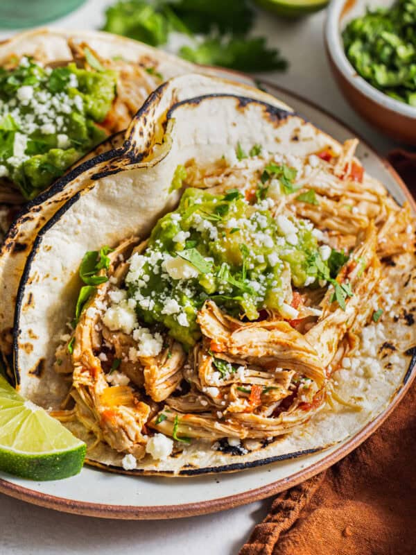 A crockpot shredded chicken taco on a plate with guacamole, cilantro, cotija cheese, and a lime wedge.